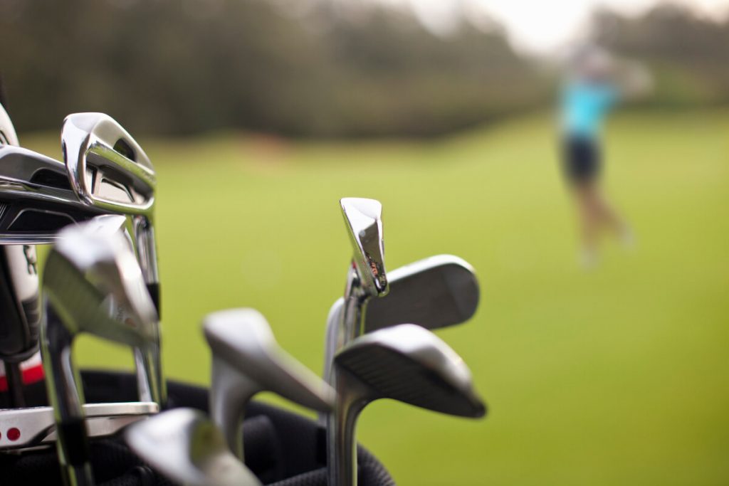 Nahaufnahme eines Golfbags mit mehreren Schlägern im Vordergrund, im Hintergrund unscharf ein Golfer beim Abschlag auf einem grünen Fairway